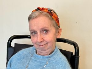 Headshot of Dr Sally Witcher. A white woman in her late 40s or so. She wears a blue wool jumper and an orange headband over short-ish light brown / grey hair. She is looking warmly into the camera.
