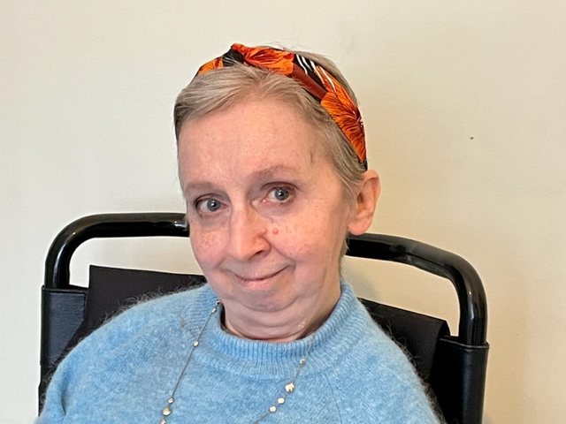 Headshot of Dr Sally Witcher. A white woman in her late 40s or so. She wears a blue wool jumper and an orange headband over short-ish light brown / grey hair. She is looking warmly into the camera.