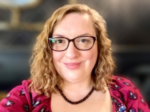 Michelle is a white female in her mid 30s. She is smiling at the camera and the picture if of her head and shoulders. She has light brown wavy hair down to her shoulders. She is wearing dark rimed glasses that much the bedded necklace she is wearing and her brown eyes. She is wear a pink top which is dotted with blue and white.