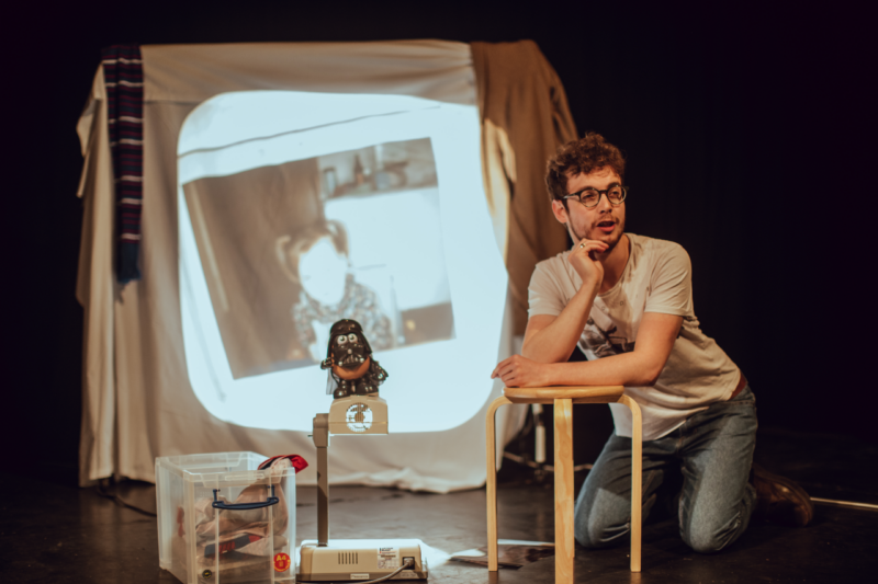 Jack kneels on the ground, leaning with his elbows on a small wooden stool. Next to him, the Drath Tater sits atop an overhead projector, which is projecting an image of Bec as a toddler sitting in a highchair, with pigtails. Jack's chin rests in his right hand as he smiles confidently away from the camera.