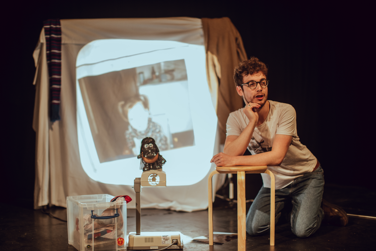 Jack kneels on the ground, leaning with his elbows on a small wooden stool. Next to him, the Drath Tater sits atop an overhead projector, which is projecting an image of Bec as a toddler sitting in a highchair, with pigtails. Jack's chin rests in his right hand as he smiles confidently away from the camera.