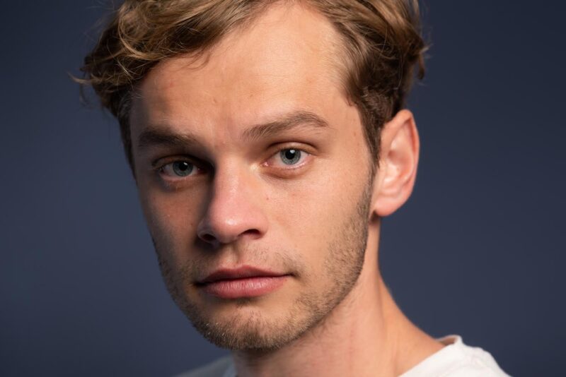 Close up shot of Aiden, a white man in his late 20s. He has short bown hair and some slight stubble coming through on his chin. He stares into the camera with a serious expression.