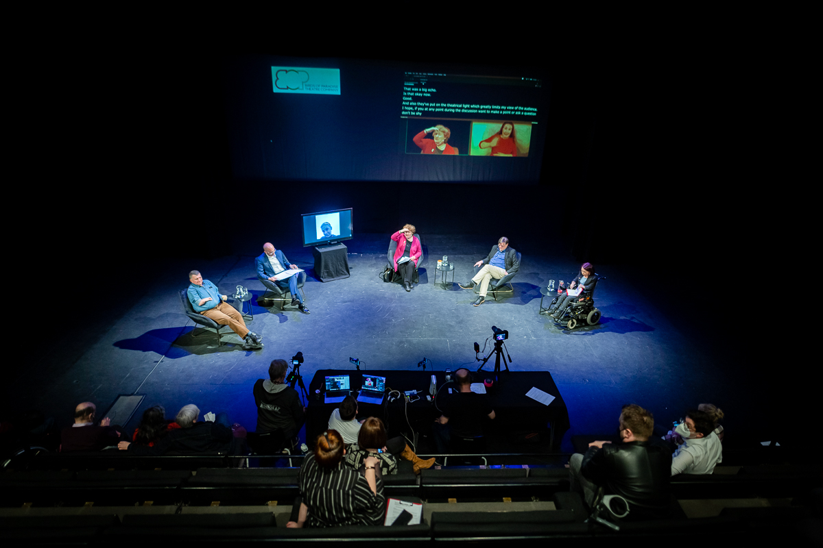 Photo of a stage from right at the back of the seating bank. It's dark with five people seated in a curve on the stage. Also in place is a large tv screen, for one person who could not attend in person. An even larger screen behind the people displays captions and a BSL interpreter.