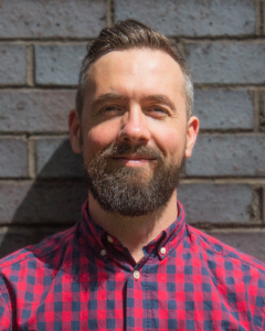 Headshot of Jamie. White man in his late 30s, short hair, with a bit longer on top and a medium sized beard. Giving a half smile to camera.