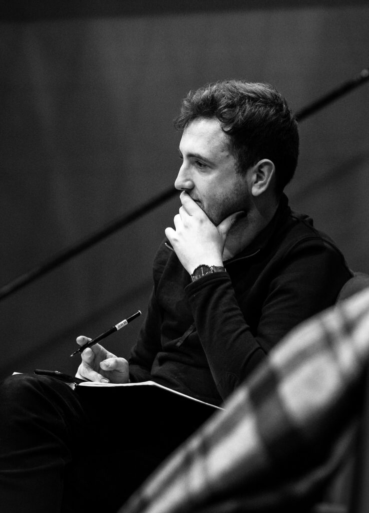Headshot of Kenneth, black and white pic of a white man in his early 30s. He sist staring off into the distance, an open note book and pen on his lap, with his left hand holding his chin in thought.