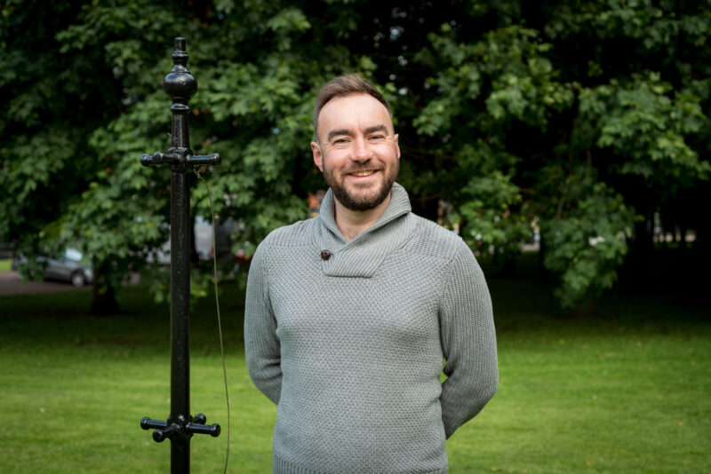 Headshot of Neil, a white man in his late 30s. He stands in a grassy area, hands clasped behind his back. He has short cropped brown hair and a beard, and a wide open smile.