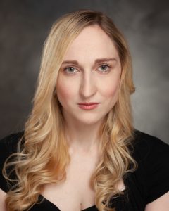 Headshot of Nicola. A white woman in her 30s, she has long curly blond hair touseling onto her chest. A serious stare into camera, with a slight smile on her lips.