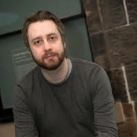 Headshot of Rob Drummond. A white man in his late 40s, with short brown hair and a beard. He looks seriously directly into camera.