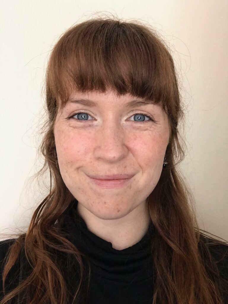 Anna is a white woman in their 20s. They have red brown hair witha fringe to just above their eyebrows and falling down past their shoulders. They are smiling warmly at the camera.