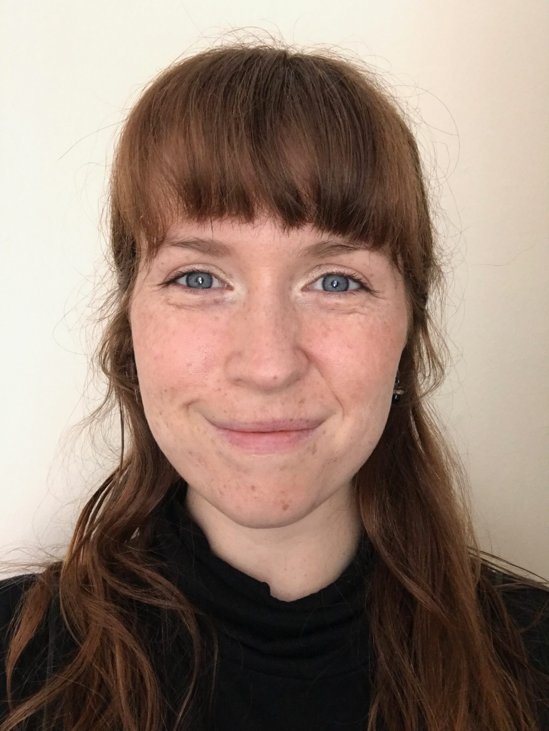 Anna is a white woman in their 20s. They have red brown hair witha fringe to just above their eyebrows and falling down past their shoulders. They are smiling warmly at the camera.