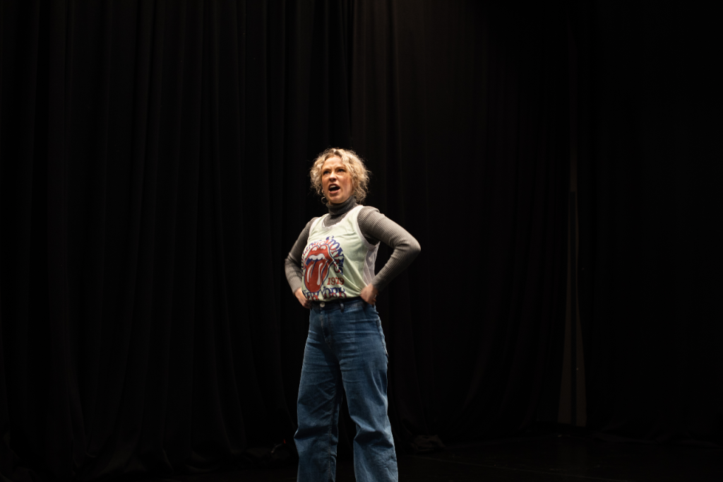 Photo of Annaleise, a white woman in her 20s with short curly blonde hair. She stands defiantly with her fists on her hips, with a stern look on her face as if telling someone off.