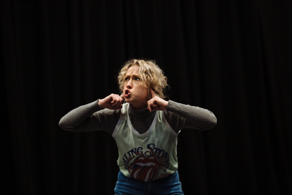 Photo of Annaliese, a white woman in her 20s with curly bonde hair down to her ears. She is stooping at the waist and has her fingers in her ears. Her lips are pouted in a "shhhh" shape.