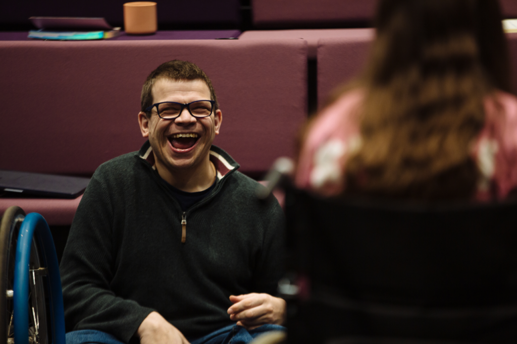 Photo of Robert, a white man in his 40s with short brown hair and glasses. He is sitting and has a wide open smile. He facing another person but we can only see their back and they are out of focus.