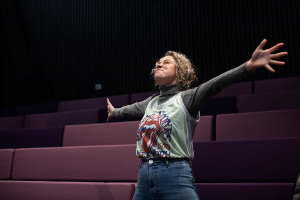 Photo of Annaliese, a white woman in her 20s with curly blonde hair. She stands in a theatre seating bank and has her arms stretched out wide, her eyes shut with an expression of contentment.