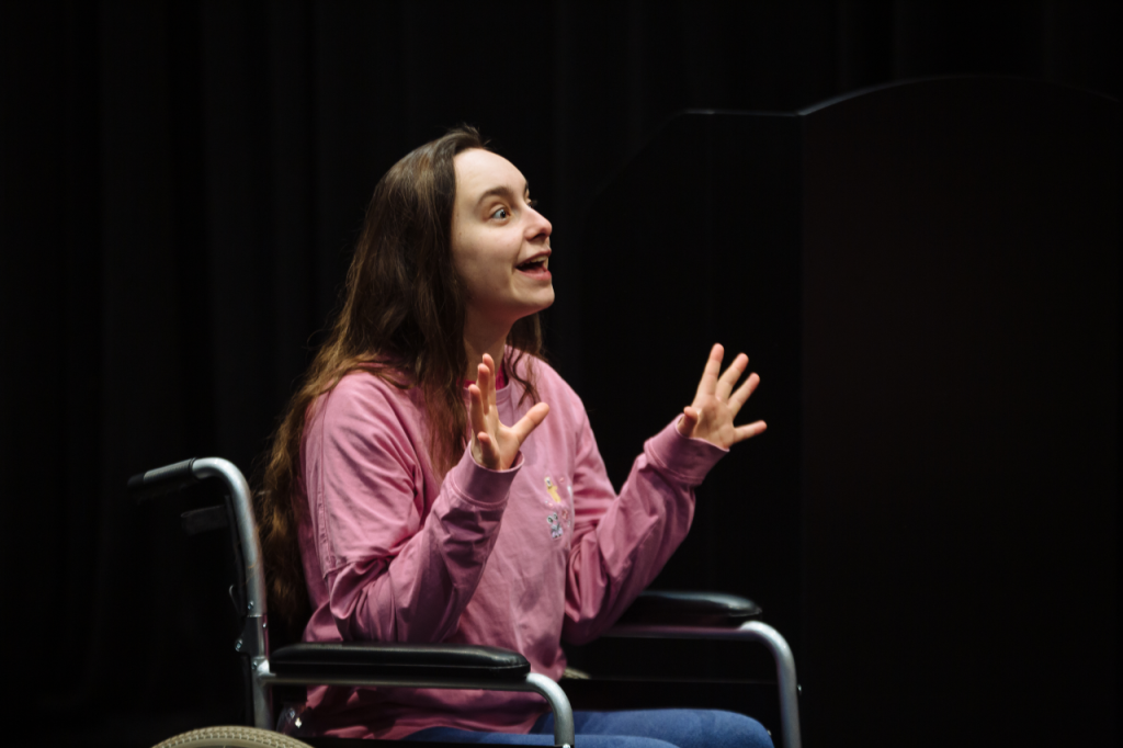 Photo of Katie, a white woman in her 20s with long brown hair. She sits in her wheelchair, with her elbows on the arms rests and the hands up in the air with the fingers splayed apart. She has a look of wonder as if she is telling an exciting story.