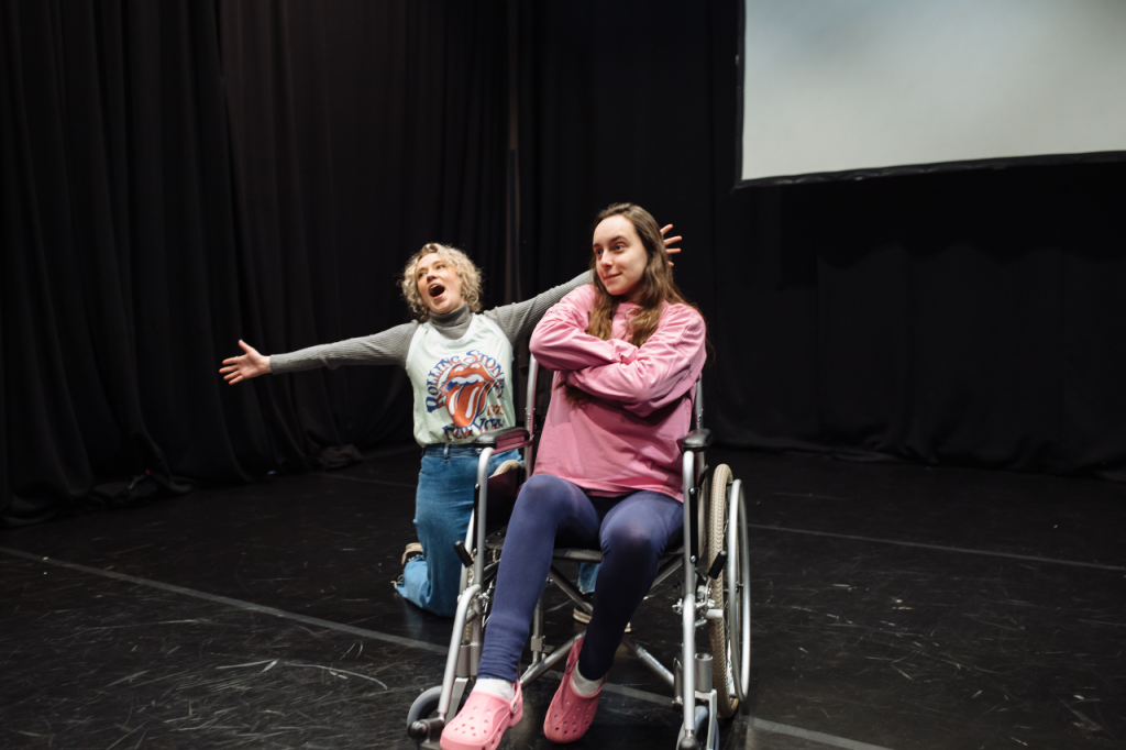 Photo of two people. In the foreground is Katie, a white woman in her 20s with long brown hair, she sits in her wheelchair with her arms crossed and held at an angle, and a smirking expression. Behind her on one knee is Annaliese, a white woman in her 20s with curly blonde hair. She has her arms open wide and her mouth open equally wide, as if in song.