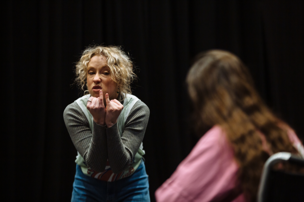 Photo of two people. On the left is Annaliese, a white woman in their 20s with curly blonde hair. They are stooping at the waist and holding both their hands up to their face, with closed fists with the little fingers on both hands extended and being held side by side. She has a bit of a sneer expression on her face. On the right, facing away from the camera, is Katie, a white woman in hers 20s with long brown hair. They are out of focus and all we can see is their left shoulder and the back of their head.