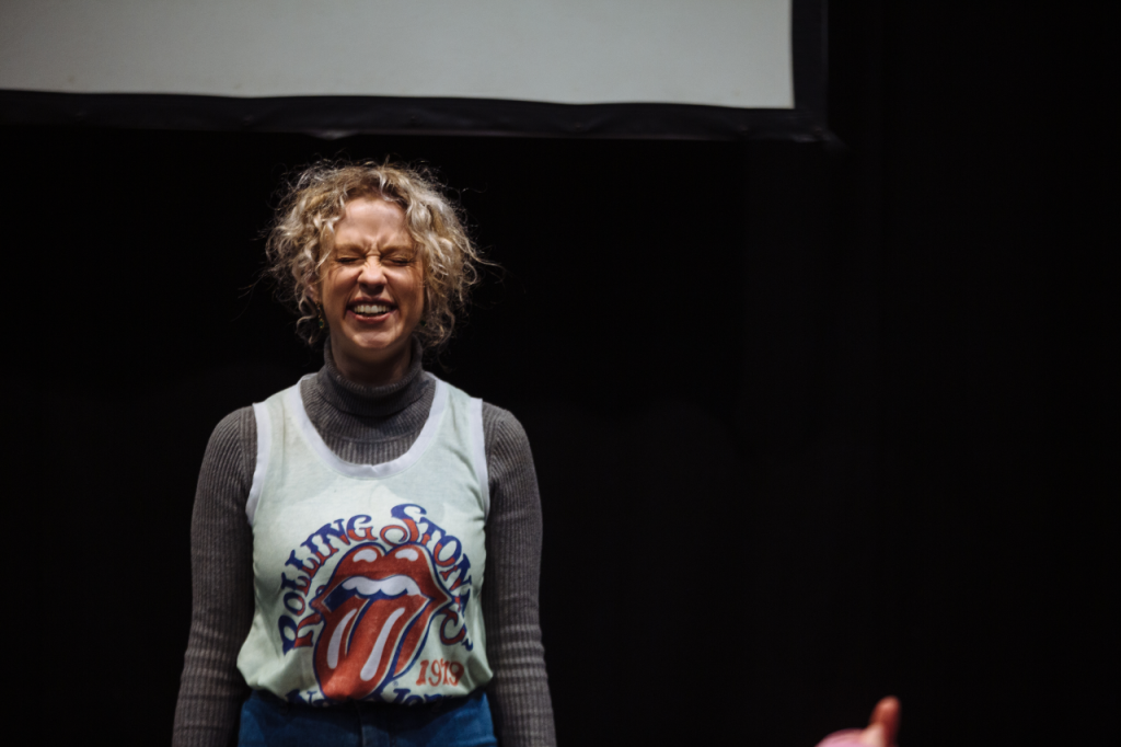 Photo of Annaliese from the waist up, a white woman in her 20s with curly blonde hair. She has her eyes screwed shut and a big grin.
