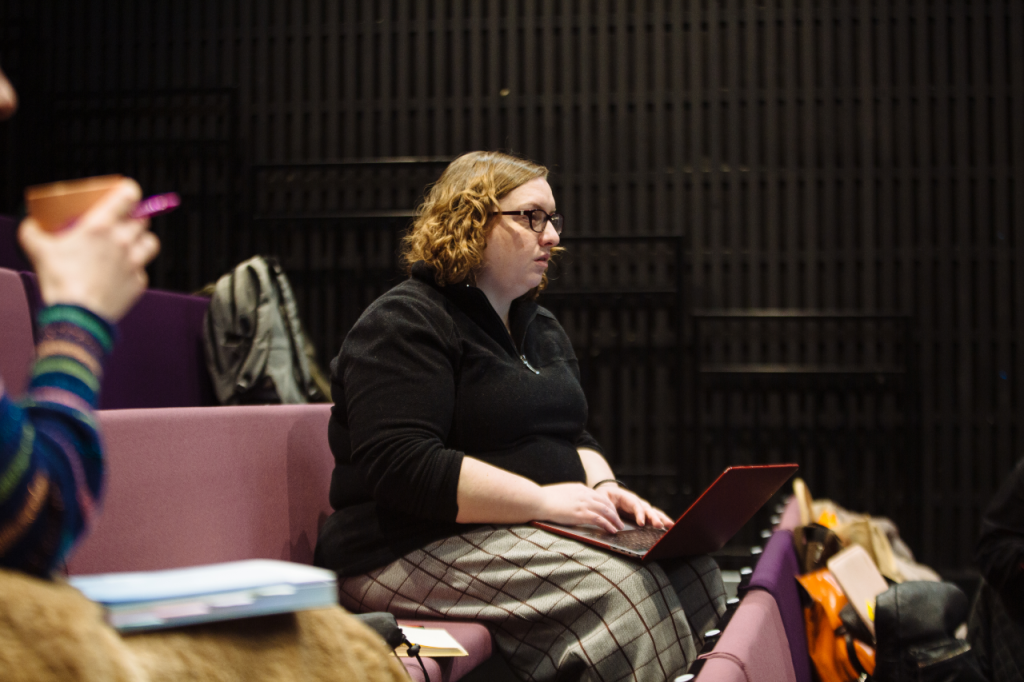 Photo of Michelle, a white woman in her 30s with shoulder length golden brown hair and glasses. She sits in a theatre seating bank with a laptop balanced on her knees. Her expression is neutral.