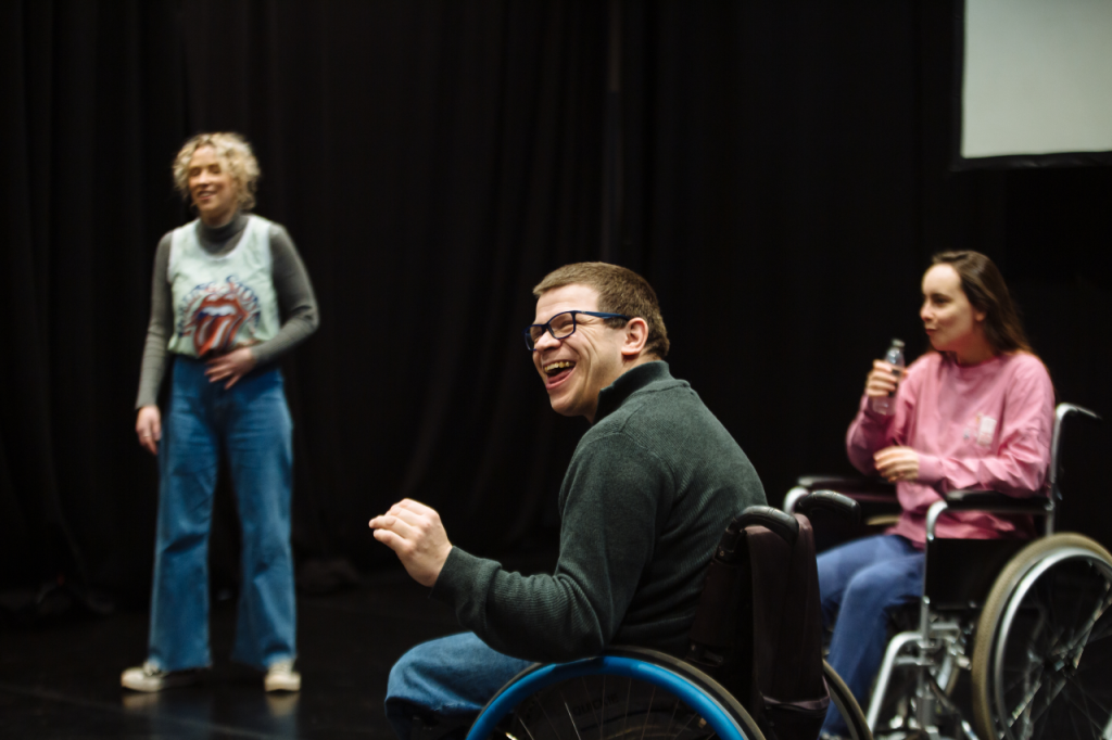 Photo of three people. In the centre foreground is Robert, a white man in his 40s with short cropped hair and glasses. He sits in his wheelchair, side on to the camera, laughing and looking at something out of shot. Standing in the background on the left is Annaliese, a white woman in her 20s with ear length curly blonde hair. Sitting in her wheelchair in the background on the right is Katie, a white woman in her 20s with long brown hair, who is swigging from a bottle.