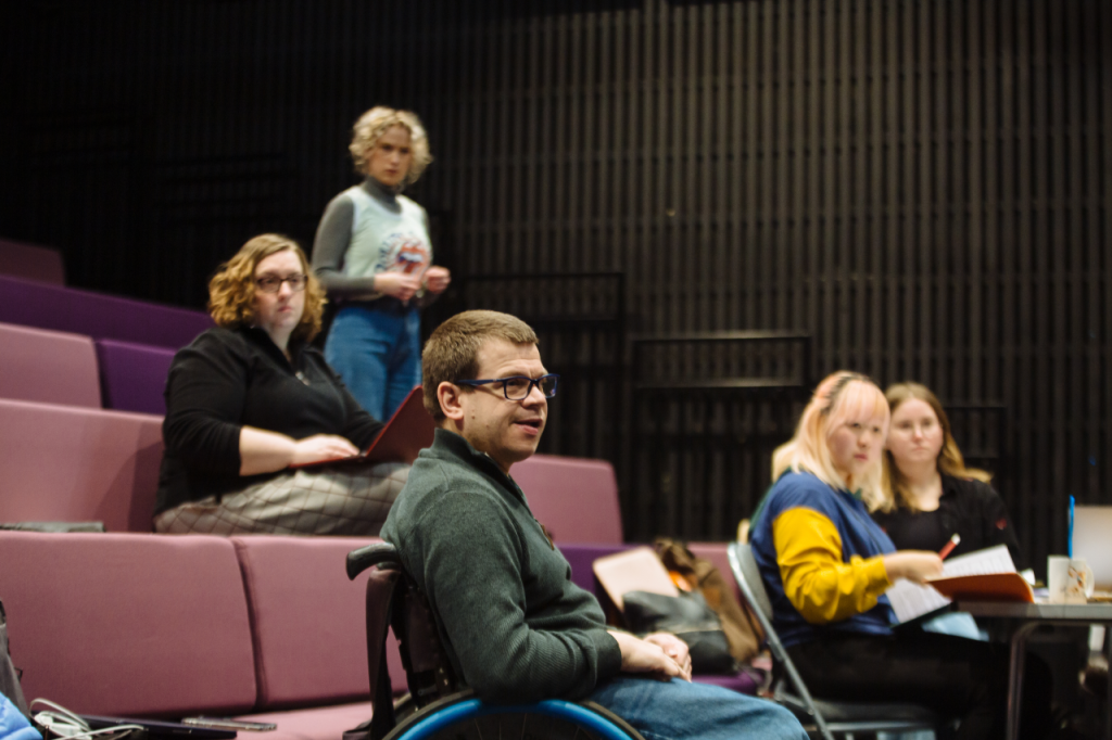 Photo of five people. In the foreground is Robert, a white man in his 40s with short brown hair and glasses. He sits in his wheelchair. Behind in in a theatre seating bank are Michelle, a white woman in her 30s, who is seated with a laptop balanced on her knee, and Annaliese, a white woman in her 20s, who is standing up. On the other side of Robert two people are seated at a table, Cynthia is a Chinese person in their 20s and Ella, a white woman in their 20s. They are all looking at something out of shot with expressions of interest.