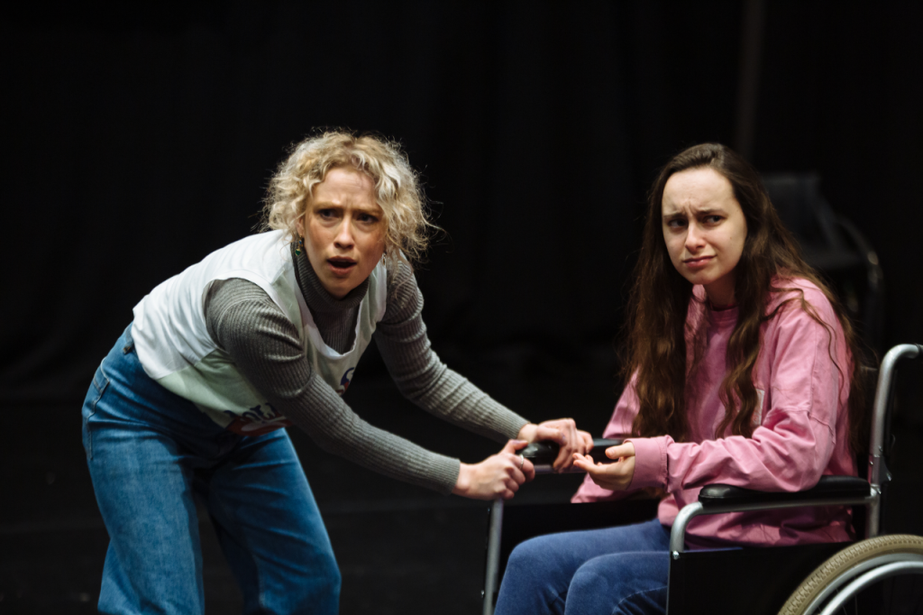 Photo of two people. On the left is Annaleise, a white woman in her early 20s with short curly blonde hair. She has a worried look on her face and is stopping over leaning over to the side with her hands grasping the arm of Katie's wheelchair. On the right is Katie, a white woman in her 20s with long brown hair. She is seated in her wheelchair and has a confused look on her face and has one hand out in front of herself with her palm facing up.