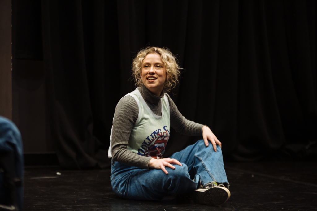 Photo of Annaliese, a white woman in her 20s with curly blonde hair down to her ears. She sits cross legged on the floor and is twisting, smiling at something out of shot.