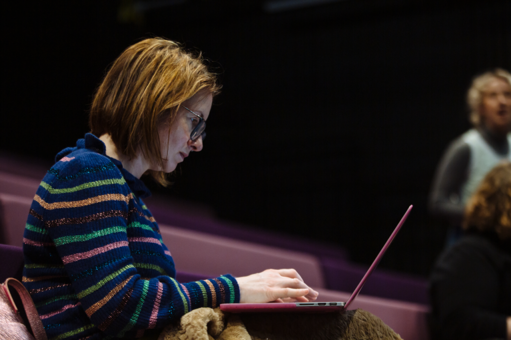 Photo of Mairi, a white woman in her 40s, with shoulder length golden brown hair. She is sitting in a theatre seating bank and is working on a laptop perched on her knees.