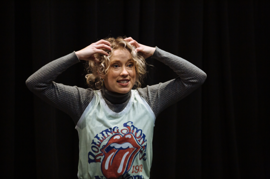 Photo of Annaliese from the waist up. She is a white woman in her 20s with curly blonde hair. She has a wide grin and both hands held to her head as if itching her scalp.