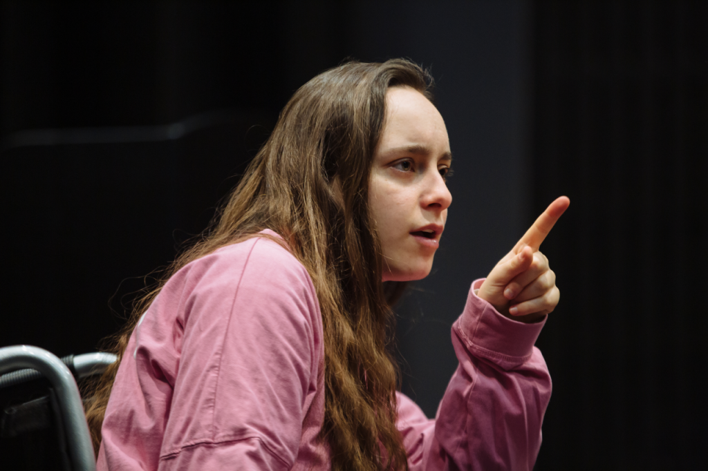 Photo of Katie from the waist up, a white woman in her 20s with long brown hair. She sits in her wheelchair with a hand raised infront of her face, with a single finger pointing upwards.