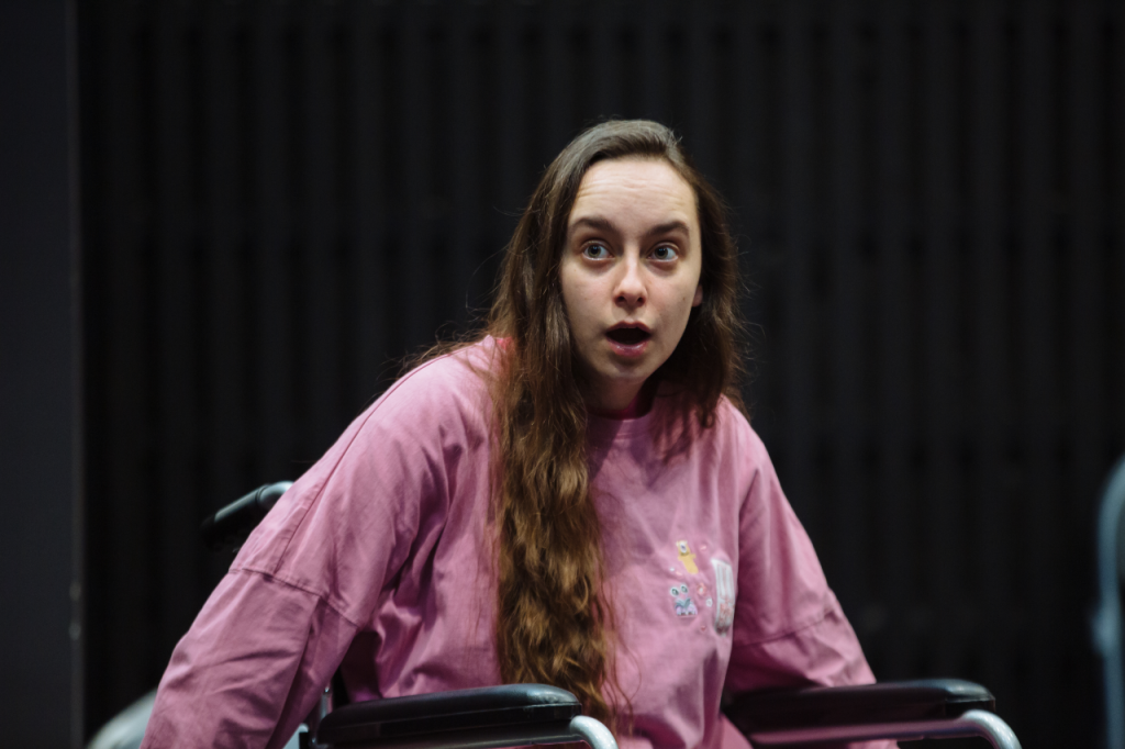 Photo of Katie from the waist up, a white woman in her 20s with long brown hair. She sits in her wheelchair with both arms extending down out of shot. She has a look of shock on her face.