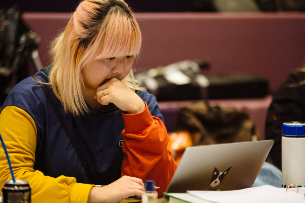Photo of Cynthia, a Chienese person in their 20s with should length blonde hair with a pink tint. She sits at a desk and concentrates on a laptop screen.