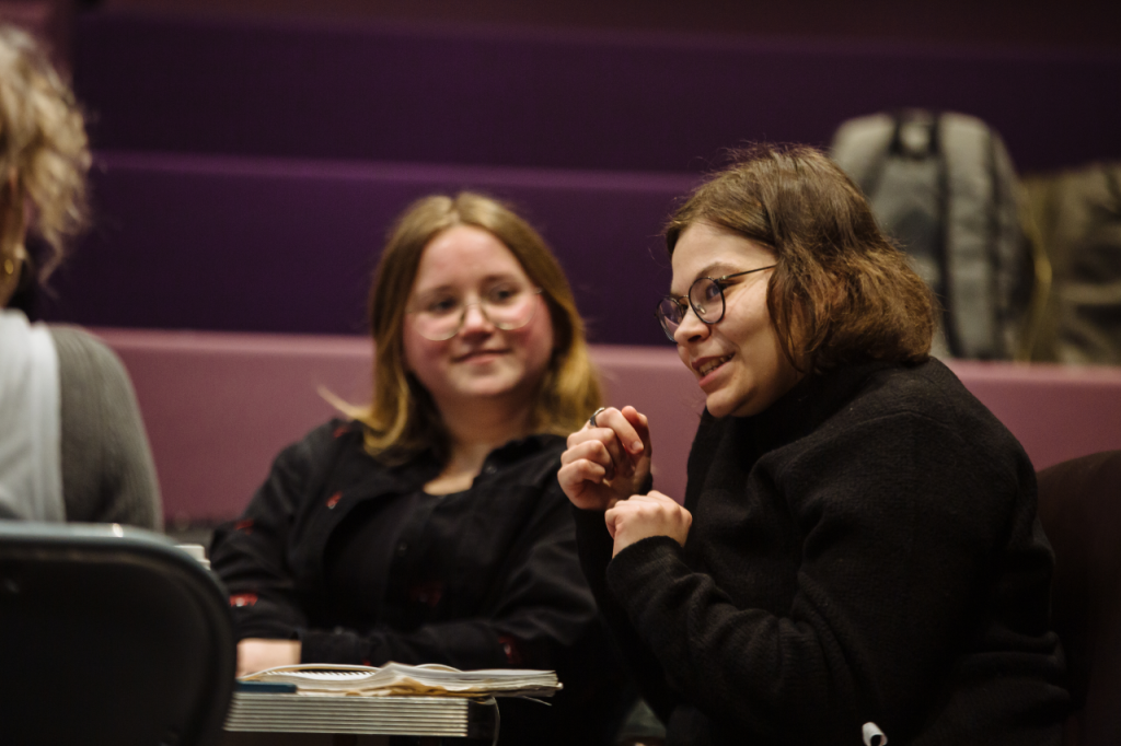 Photo of two people. Finley is a white person in their 20s with shoulder length brown hair and glasses. Ella is a white woman in her 20s with shoulder length golden brown hair, wearing glasses. They are both seated as a table and appear to be in conversation.