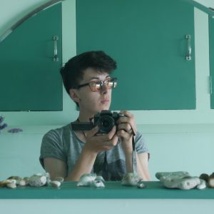 Kerry reflection is framed in a mirror. Behind them is a green wall. Kerry is white and has short black hair and is wearing glasses. They have a grey t-shirt on and is holding a black camera.