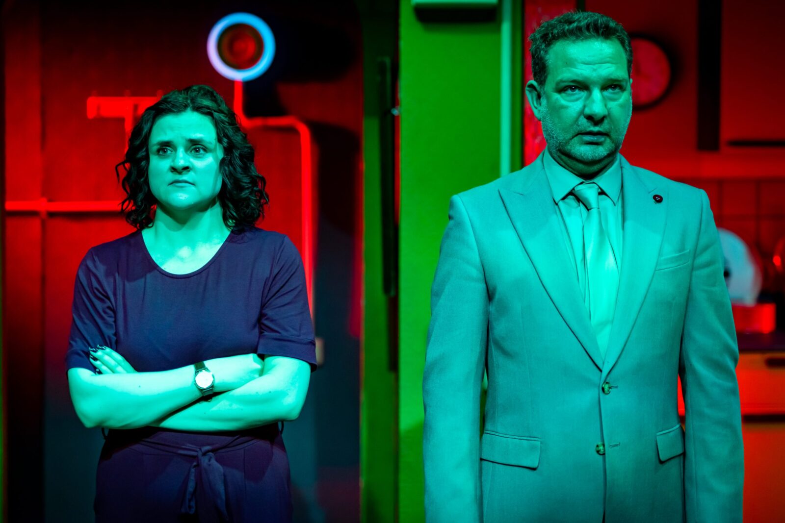 Two people standing side by side, staring straight ahead with blank expressions. On the left is a young woman with dark curly hair and wearing a purple jumpsuit. On the right is a middle aged man in a mint green suit.