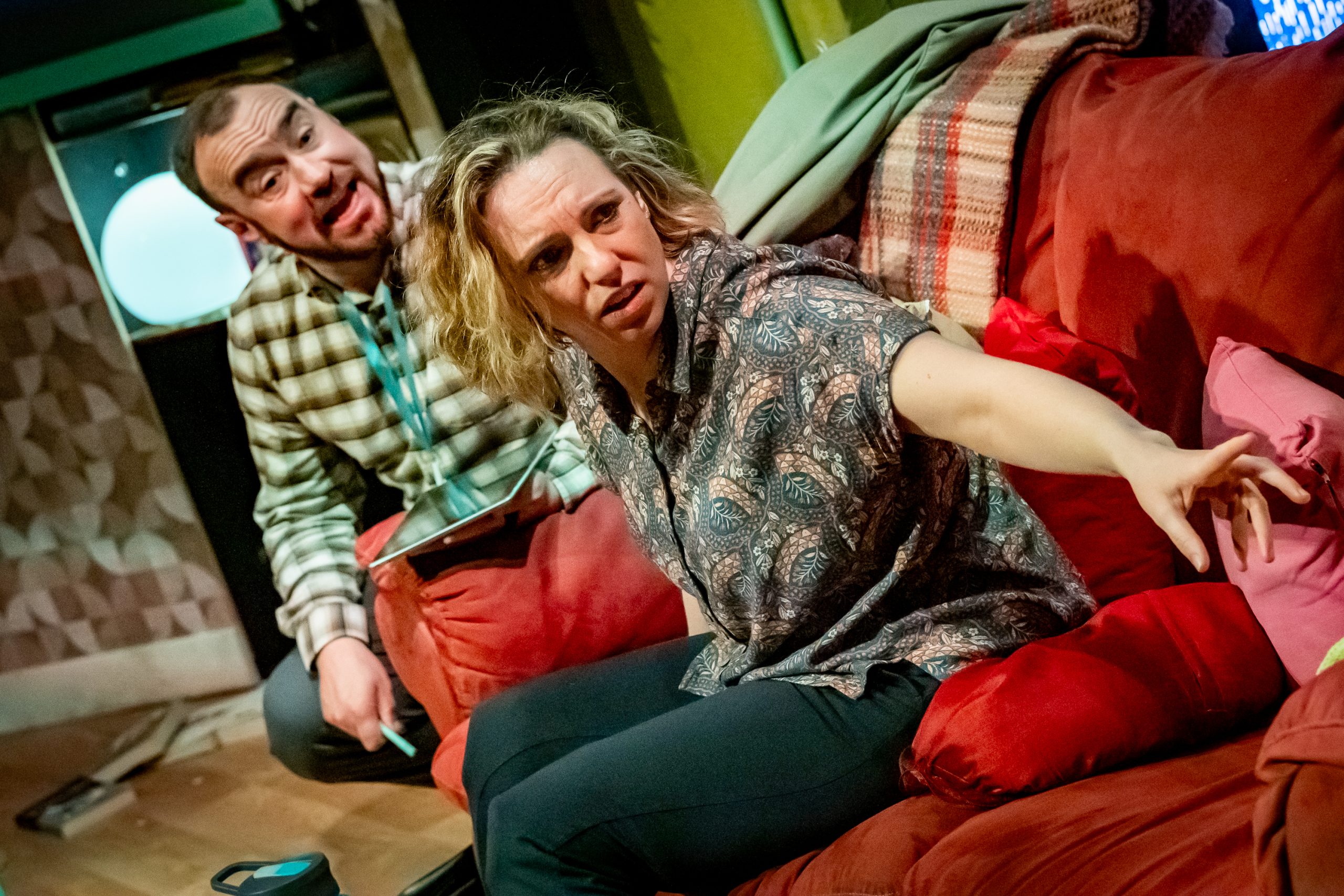 A blonde woman and a man wearing a lanyard sit on a red sofa. The woman is turning away from the man with an expression of distress, while the man attempts to reason with her.