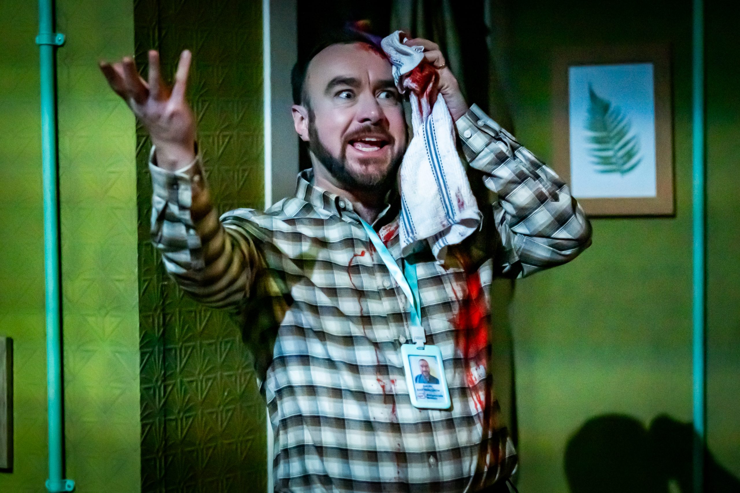 A dark-haired man with a short beard holds a tea towel to the side of his head as he speaks emphatically. The tea towel and his shirt are both stained with blood.
