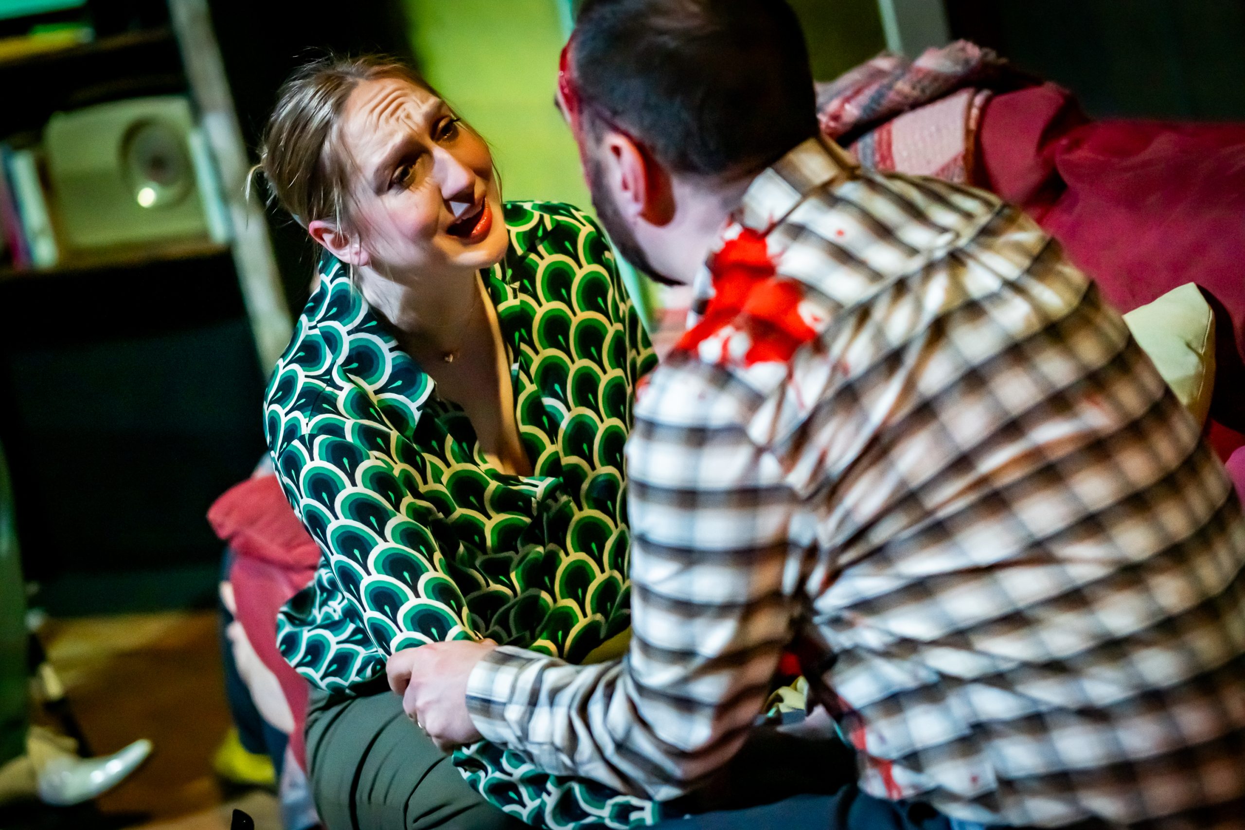 A blonde woman and a dark-haired man on a couch. The woman is holding the man's hands and pleading with him. The man is facing away from the camera. His shirt and the side of his head are covered in blood.
