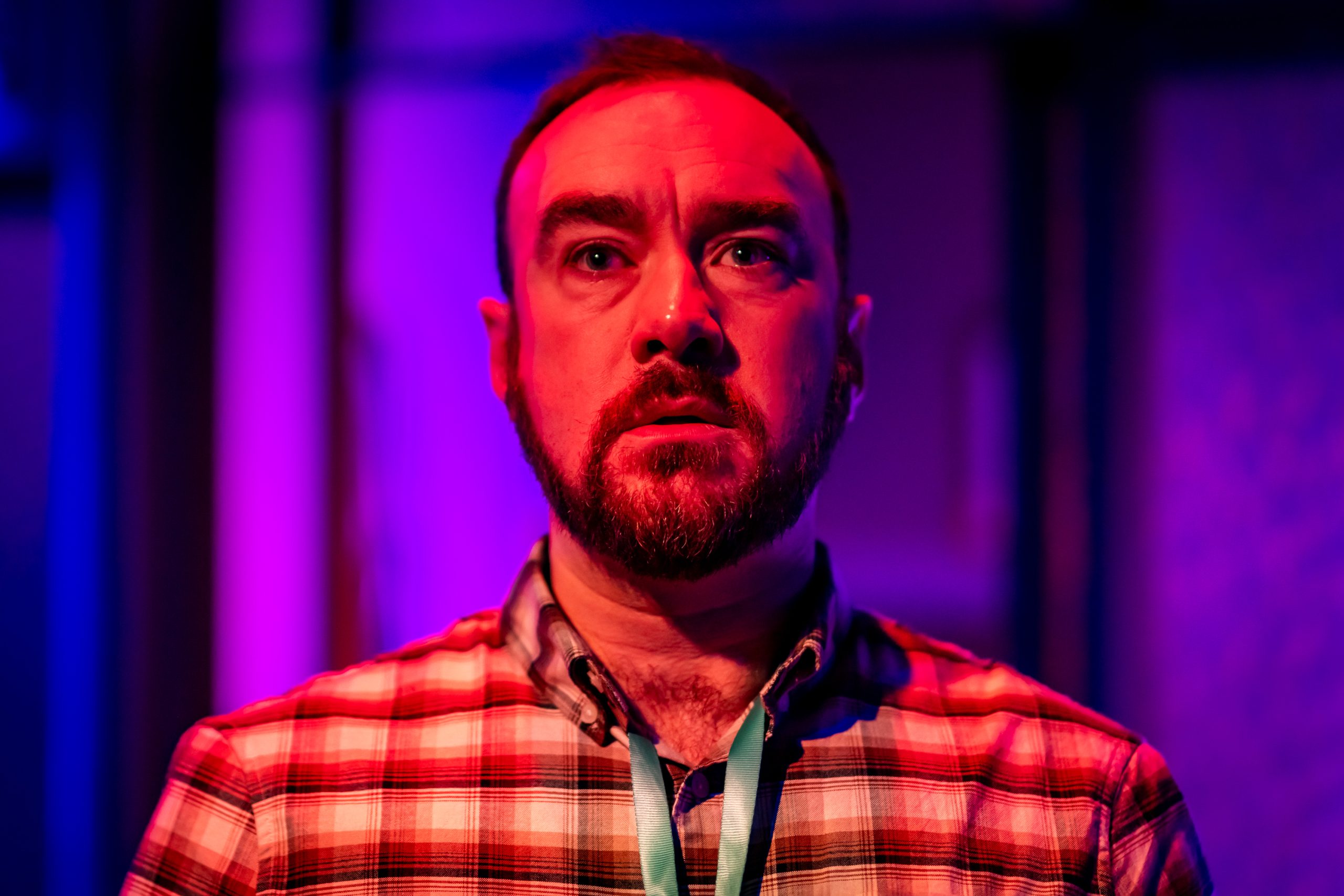 A man with short dark hair and a dark beard, wearing a checked shirt and a lanyard, stares blankly towards the camera. He is illuminated by an unnatural red light.