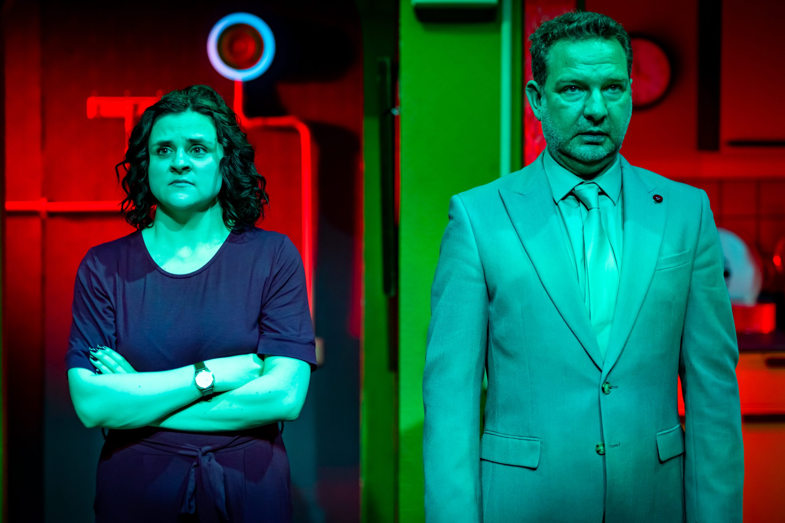 Two people standing side by side, staring straight ahead with blank expressions. On the left is a young woman with dark curly hair and wearing a purple jumpsuit. On the right is a middle aged man in a mint green suit.
