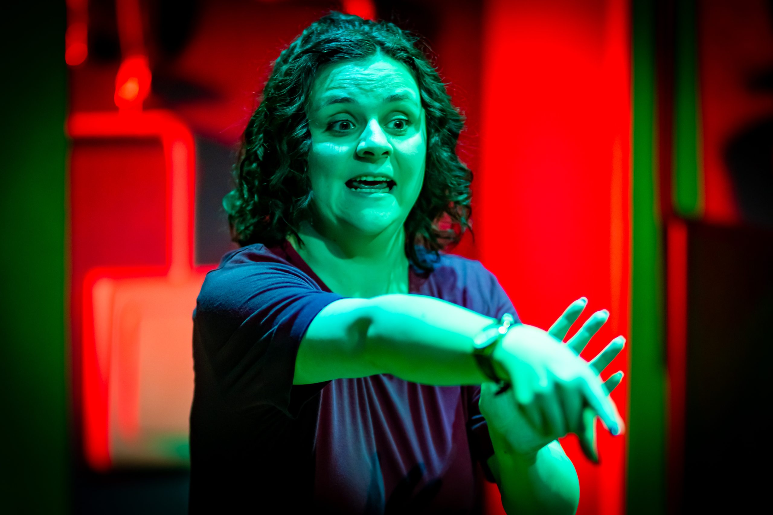 A young woman with dark curly hair and wearing a purple jumpsuit uses BSL. She is illuminated by an unnatural green light and pointing at something out of frame.