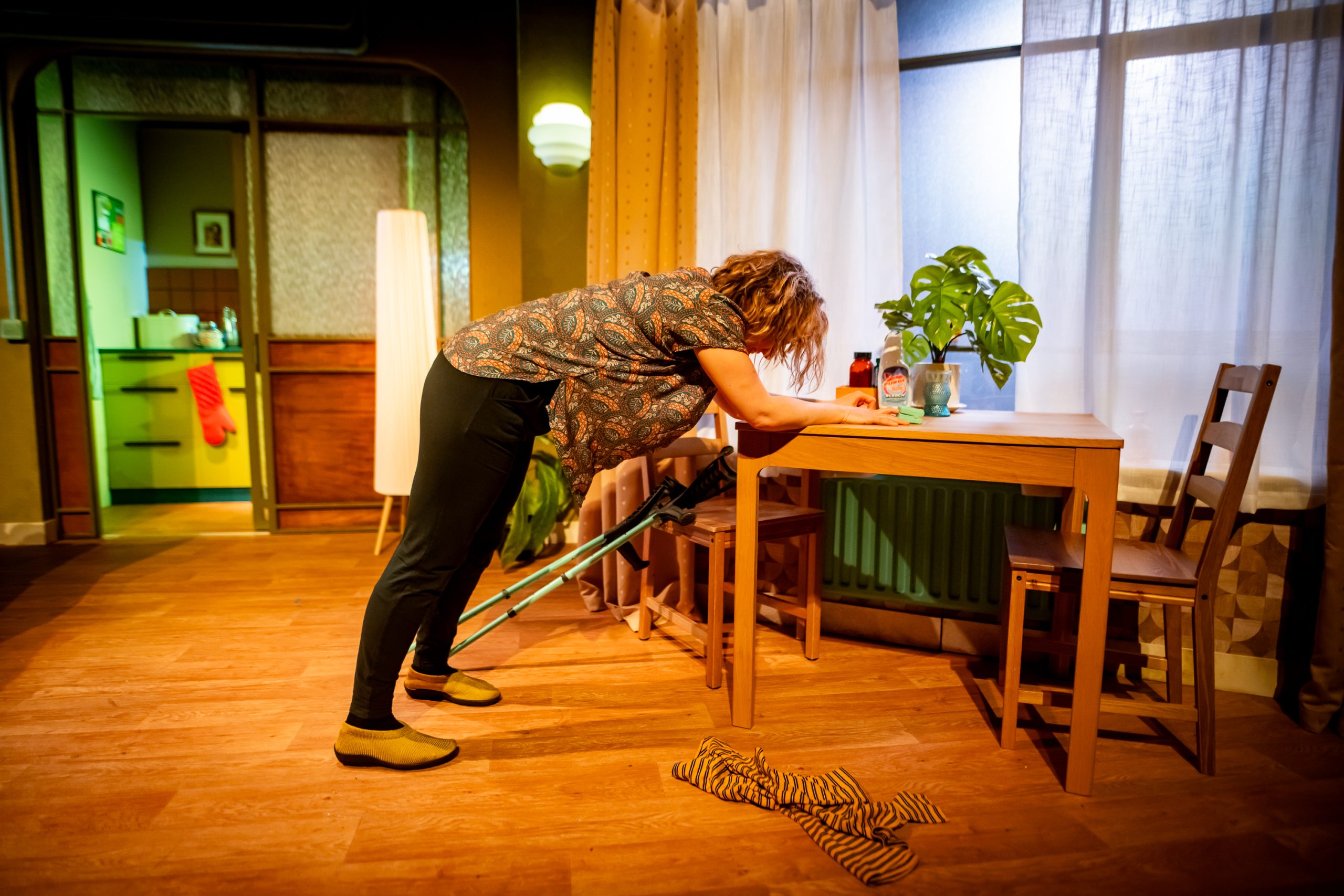 A blonde woman stretches her legs and back by leaning on a dining table. Two mint green forearm crutches are balanced against the table next to her.