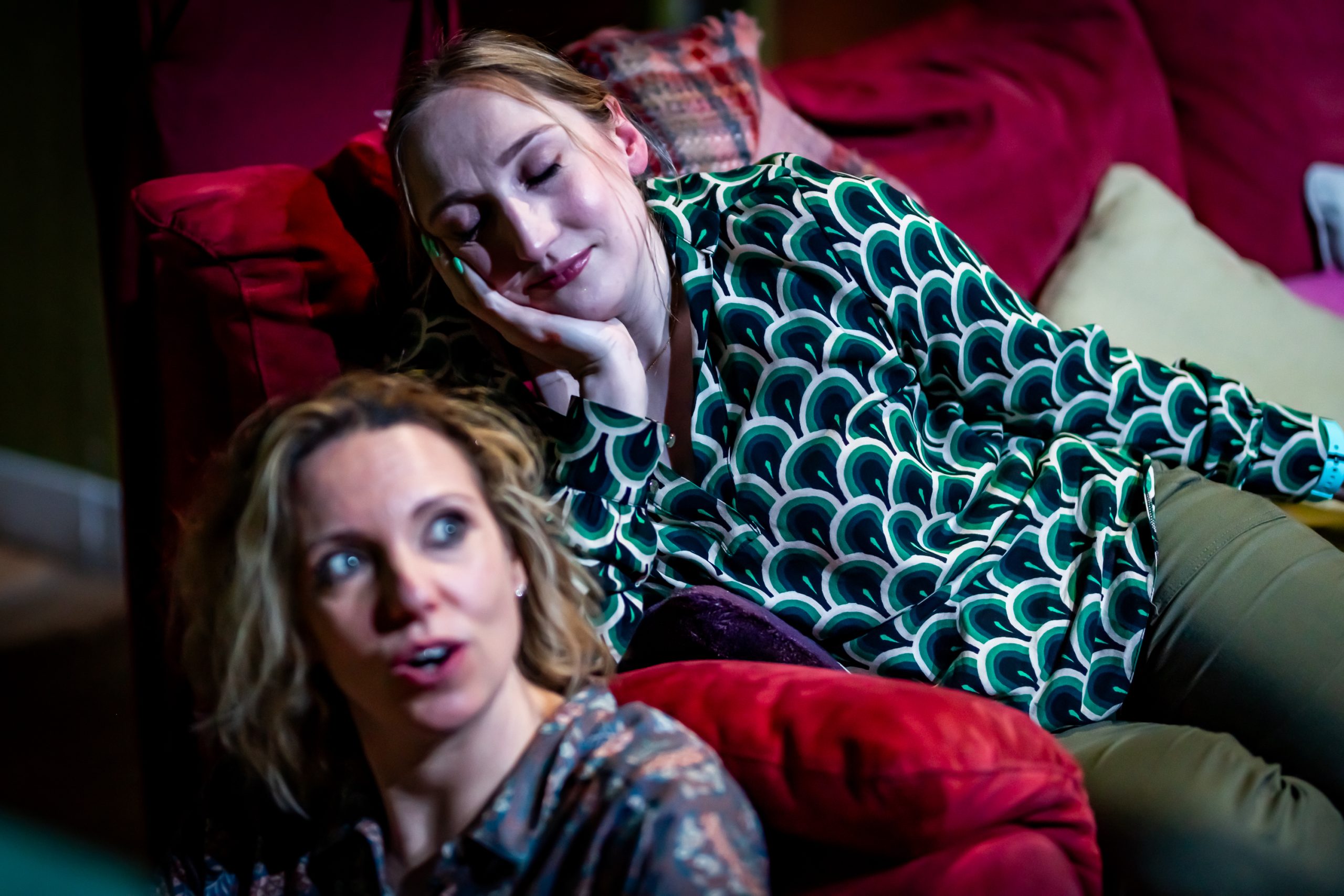 A woman with a blonde ponytail and wearing a shiny top leans on the arm of a sofa, looking serene. A woman with shoulder-length hair is sitting on the floor against the sofa, speaking to the first woman without looking at her.