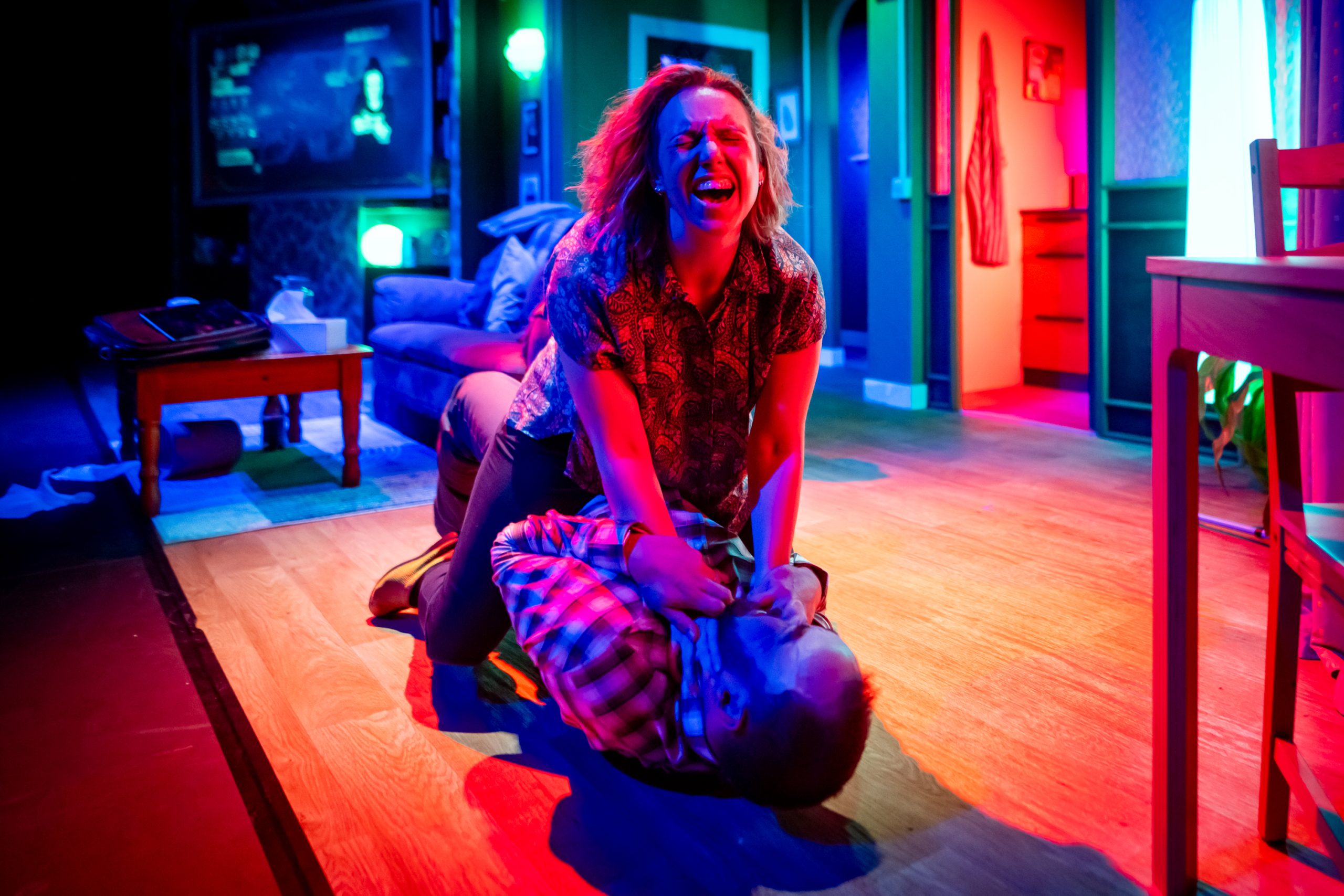 A blonde woman strangles a dark-haired man on the floor of a living room. She has an expression of anguish as he struggles.