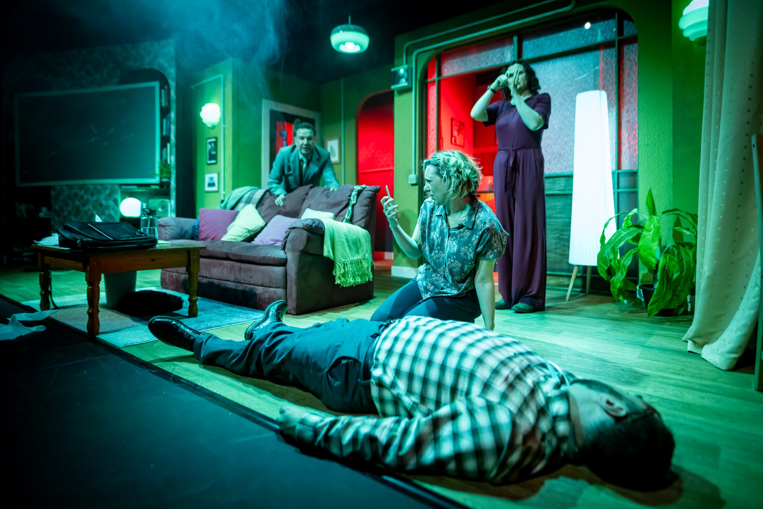 A neatly dressed man lies unresponsive on the floor of a living room set. A blonde woman is sitting next to him and squinting at a cellphone in her hand. A man in a mint suit and a woman in a purple jumpsuit look horrified in the background.