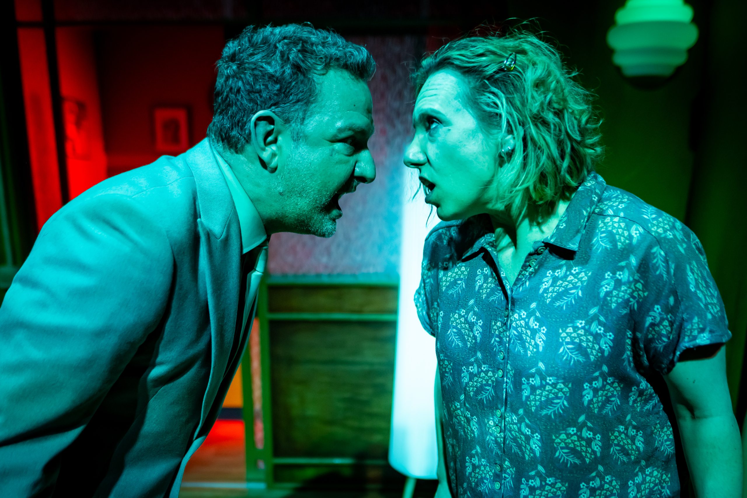 A woman with shoulder-length blonde hair and a middle-aged man in a mint green suit face each other. Both are yelling and their faces are nearly touching. They are illuminated by an unnatural teal light.