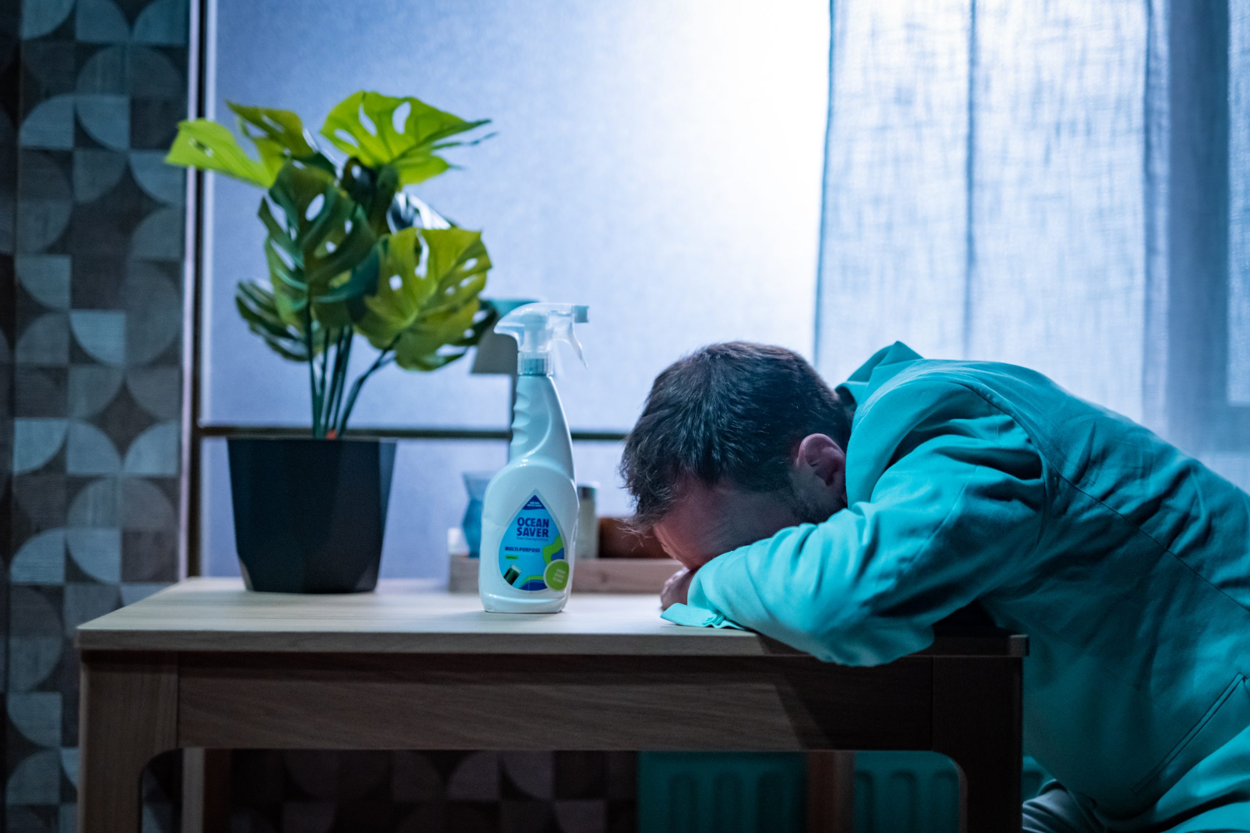 A dark-haired man in a mint green suit sits at a dining table with his head against the table. A spray bottle of bleach and a potted plant sit next to him.