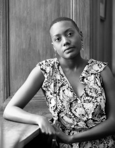 A young woman with short cropped hair, long earrings and a floral top, leaning on a bench and looking at the camera with a serious expression.