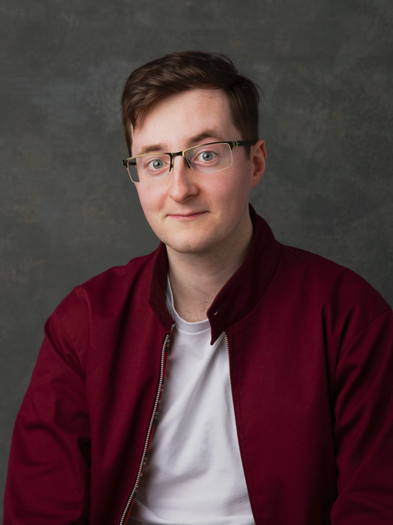 A young man with short hair and glasses, wearing a white tshirt and maroon jacket, with a slight smile.
