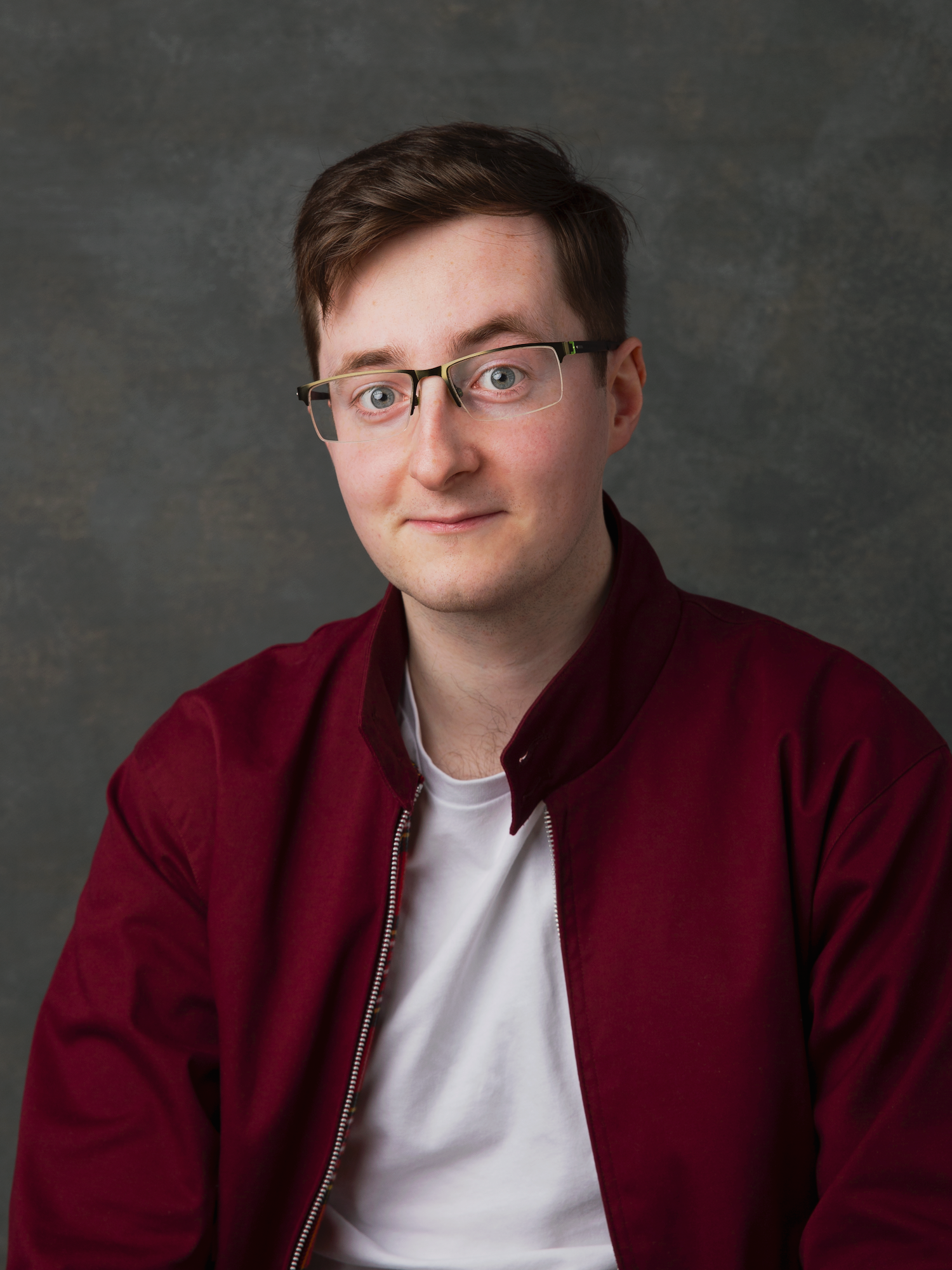 A young man with short hair and glasses, wearing a white tshirt and maroon jacket, with a slight smile.